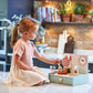 A young girl sits on a kitchen counter playing with the Tender Leaf Toys Kitchenette, holding a wooden spatula over a pot and surrounded by wooden utensils.