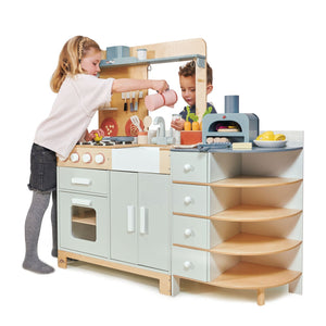 Two children are playing with the Tenderleaf La Fiamma Grand Kitchen by Tender Leaf Toys. The girl is pouring into a pot, and the boy is washing vegetables. The kitchen set includes an oven, sink, stove, and shelves with various accessories to enhance their fun.