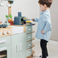 A young boy in a blue shirt plays with the Tenderleaf La Fiamma Grand Kitchen from Tender Leaf Toys, using a toy spatula to move a piece of pretend food near the mini pizza oven and other fun accessories.