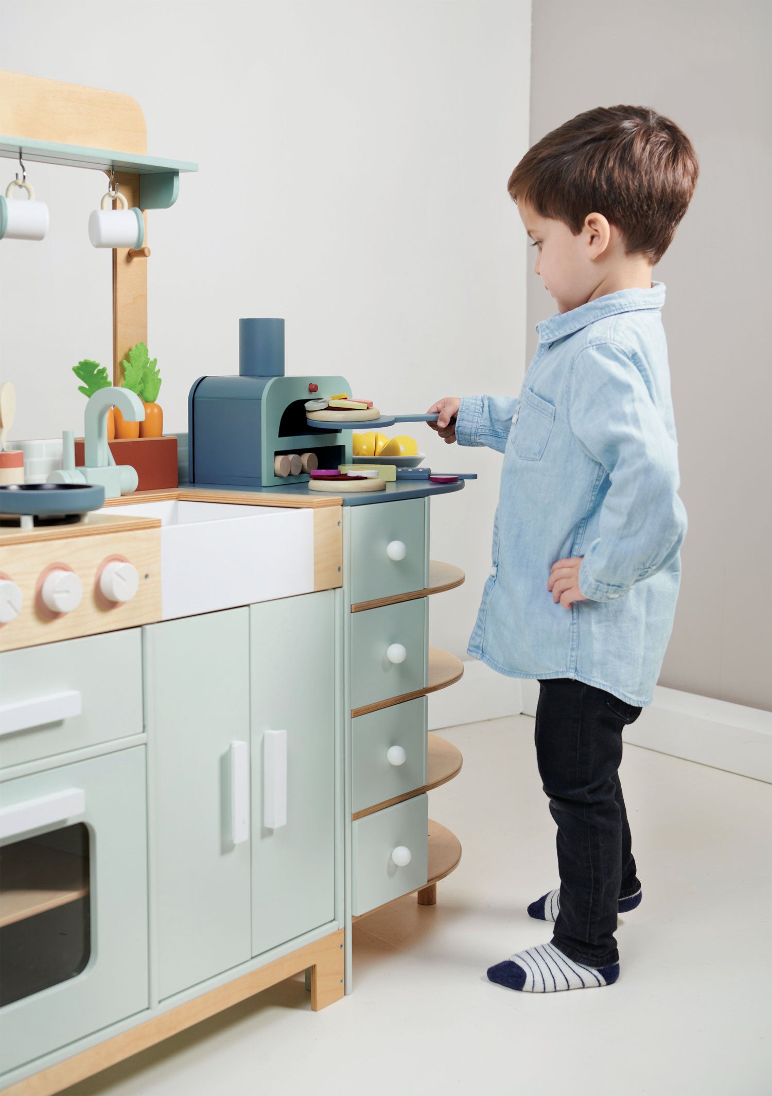 A young boy in a blue shirt plays with the Tenderleaf La Fiamma Grand Kitchen from Tender Leaf Toys, using a toy spatula to move a piece of pretend food near the mini pizza oven and other fun accessories.