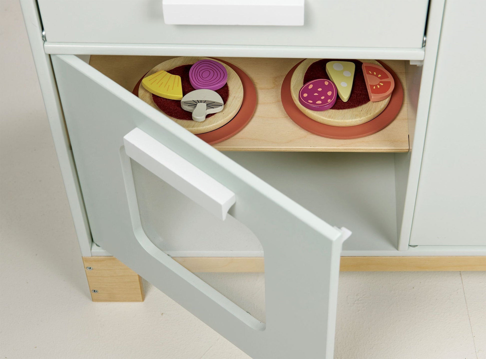 A light green cabinet door is open, revealing two wooden toy pies placed on a shelf inside. The pies, resembling creations from the Tenderleaf La Fiamma Grand Kitchen by Tender Leaf Toys, are topped with various toy vegetables and accessories.