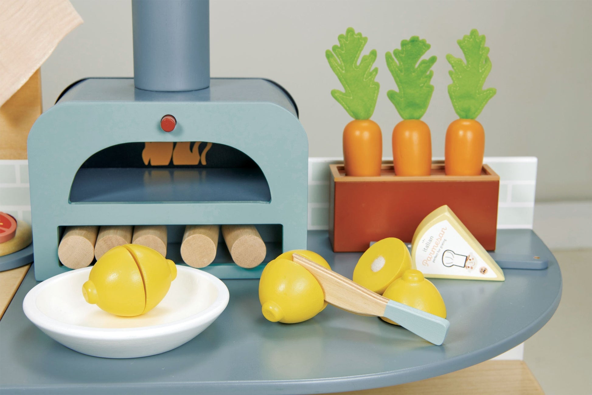 The Tenderleaf La Fiamma Grand Kitchen by Tender Leaf Toys features a toy kitchen with wooden lemons in a bowl, toy carrots in a planter, a triangular food piece, and wooden logs. Complete with a pretend cooking stove and pizza oven, this set has all the accessories your little chef needs for imaginative play.