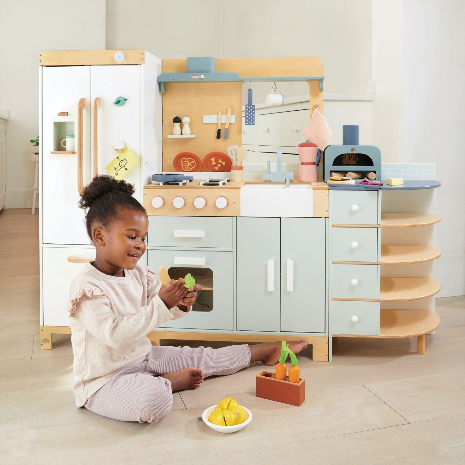 A child plays with toy food in front of the Tenderleaf La Fiamma Grand Kitchen by Tender Leaf Toys, which includes a refrigerator, stove, sink, pizza oven, and shelves stocked with accessories.