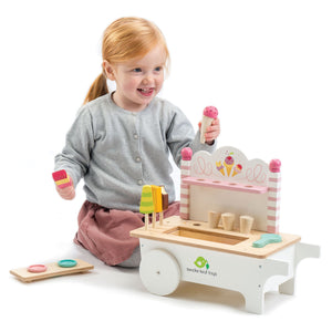 A young child engages in imaginative play with the Tender Leaf Toys Ice Cream Cart, holding a pretend ice cream cone and treat, while wooden accessories from the set are scattered on the floor nearby.