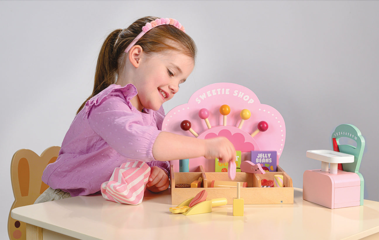 A young girl in a purple shirt plays with the Tender Leaf Toys Sweetie Pic'n' Mix Shop at a white table, using tongs and pretend pic n mix candies.