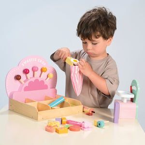 A young child places a toy candy into a striped bag at the Tender Leaf Toys Sweetie Pic'n' Mix Shop, with colorful pretend candies displayed on the wooden table, capturing the nostalgic charm of a retro sweet shop.