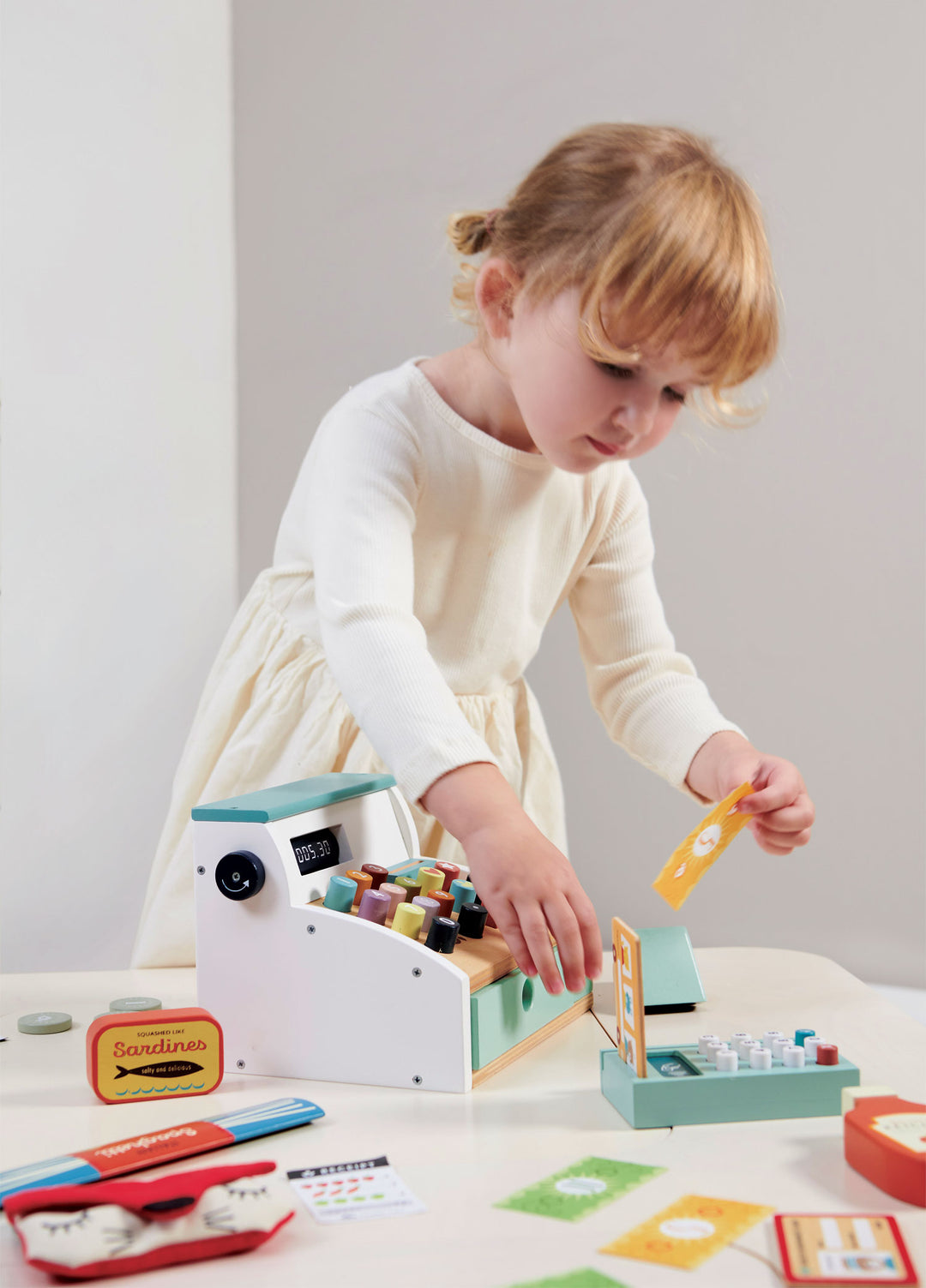 A child in a white dress gleefully plays with the Tenderleaf General Stores Till by Tender Leaf Toys on a table, swiping an imaginary credit card through the playful wooden cash register.