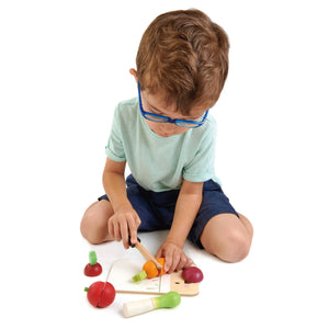 A young boy wearing blue glasses sits on the floor and plays with the Tender Leaf Toys Mini Chef Chopping Board, using its wooden knife and sustainable rubber wood chopping board to cut toy vegetables.