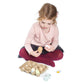 A young child sits cross-legged, holding two Tender Leaf Toys Wooden Eggs, with an open toy egg carton and several wooden eggs and pieces arranged on the floor in front of her, fully immersed in pretend play.