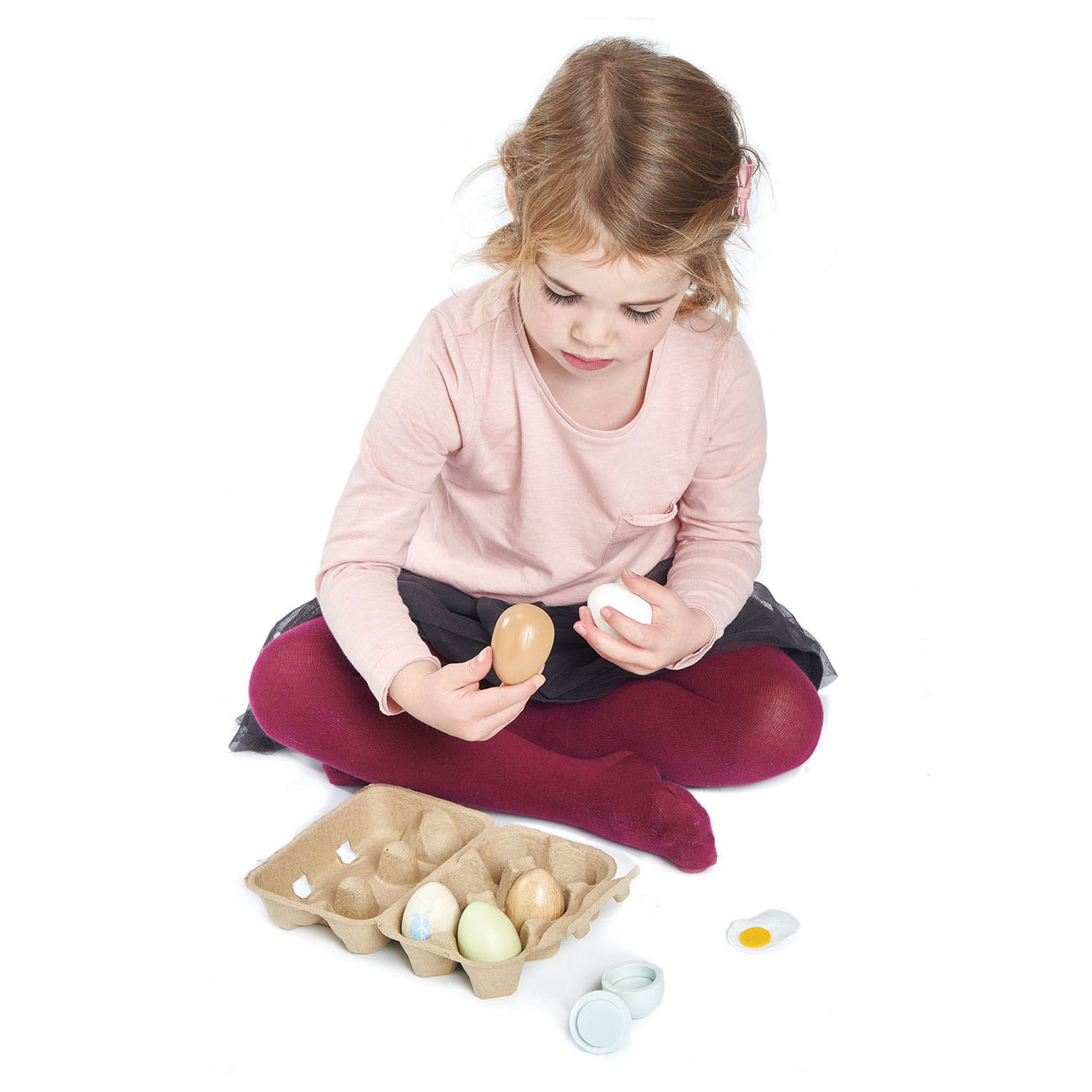 A young child sits cross-legged, holding two Tender Leaf Toys Wooden Eggs, with an open toy egg carton and several wooden eggs and pieces arranged on the floor in front of her, fully immersed in pretend play.