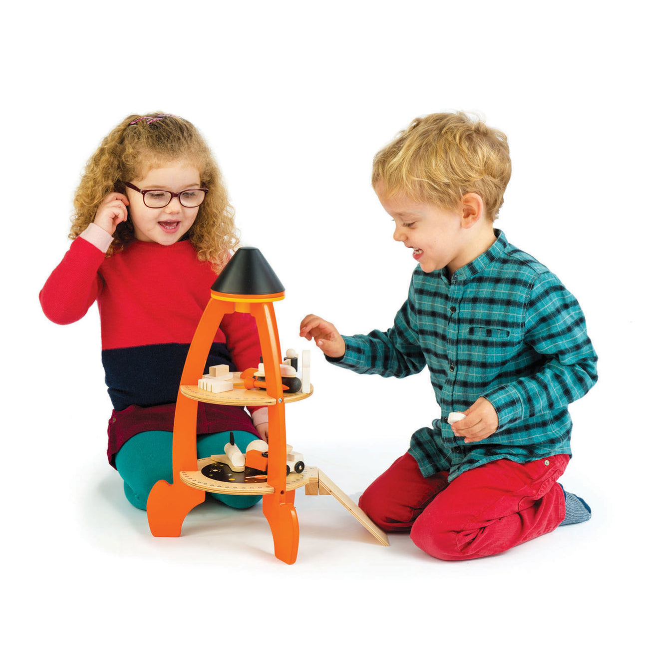 Two young children sit on the floor playing with the vibrant Tender Leaf Toys Cosmic Rocket Set, crafted from orange and natural wood. The girl wears glasses and a red dress, while the boy sports a blue plaid shirt and red pants.
