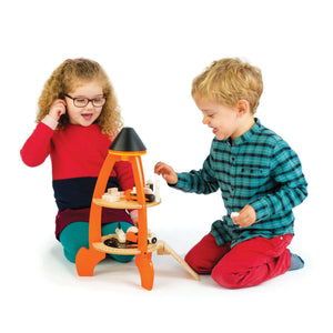 Two young children sit on the floor playing with the vibrant Tender Leaf Toys Cosmic Rocket Set, crafted from orange and natural wood. The girl wears glasses and a red dress, while the boy sports a blue plaid shirt and red pants.
