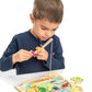 A young boy uses the magnetic fishing rod from his Tender Leaf Toys Pond Dipping set to pick up pieces from the wooden fishing puzzle game on a white table, helping develop his fine motor skills.