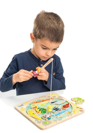 A young boy uses the magnetic fishing rod from his Tender Leaf Toys Pond Dipping set to pick up pieces from the wooden fishing puzzle game on a white table, helping develop his fine motor skills.