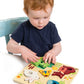 A toddler sits on a light blue chair, playing with the Tender Leaf Toys Touchy Feely Animals puzzle by placing a green frog piece onto the board and enjoying tactile learning.