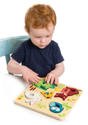 A toddler sits on a light blue chair, playing with the Tender Leaf Toys Touchy Feely Animals puzzle by placing a green frog piece onto the board and enjoying tactile learning.