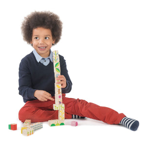 A child with curly hair, wearing a navy sweater and red pants, sits on the floor and builds a tall tower with Tender Leaf Toys Garden Blocks, developing hand-eye coordination as they carefully balance each piece.