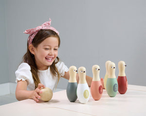 A young girl smiles as she prepares to roll a wooden ball toward a row of six colorful wooden skittles from her vibrant Birdie Skittles game by Tender Leaf Toys on the table.