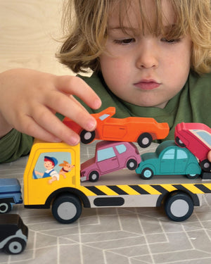 A child in a green shirt sits at a table, stacking the colorful Tender Leaf Toys Stacking Cars onto a yellow toy truck, enjoying a fun game that helps develop fine motor skills.