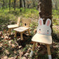 Three Tender Leaf Toys Forest Rabbit Chairs sit outdoors on grass and leaves near a tree in a sunlit forest.