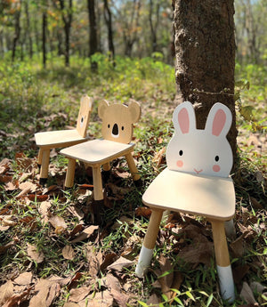 Three Tender Leaf Toys Forest Rabbit Chairs sit outdoors on grass and leaves near a tree in a sunlit forest.