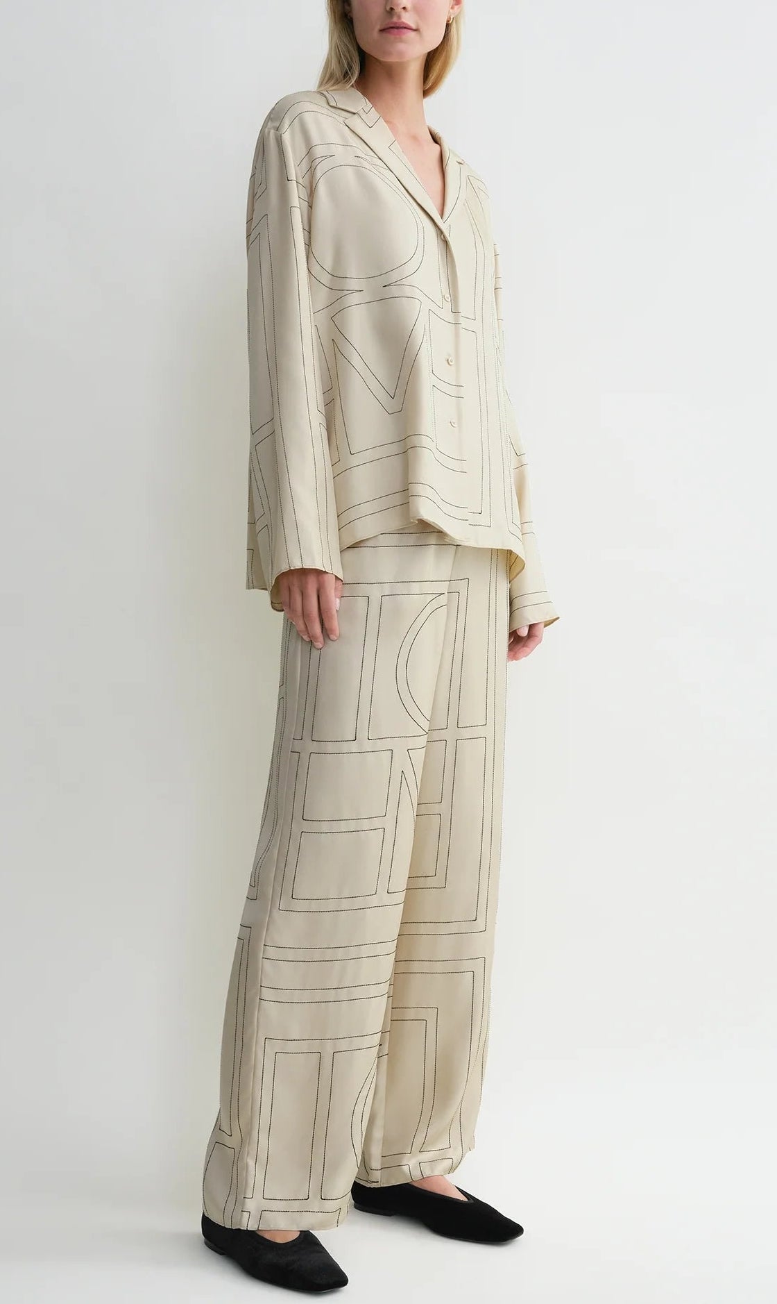 A person wearing a loose-fitting, cream-colored Toteme Monogram Silk PJ Top with geometric line patterns, standing against a plain white background.