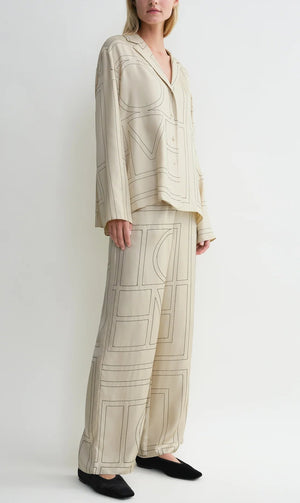 A person wearing a loose-fitting, cream-colored Toteme Monogram Silk PJ Top with geometric line patterns, standing against a plain white background.