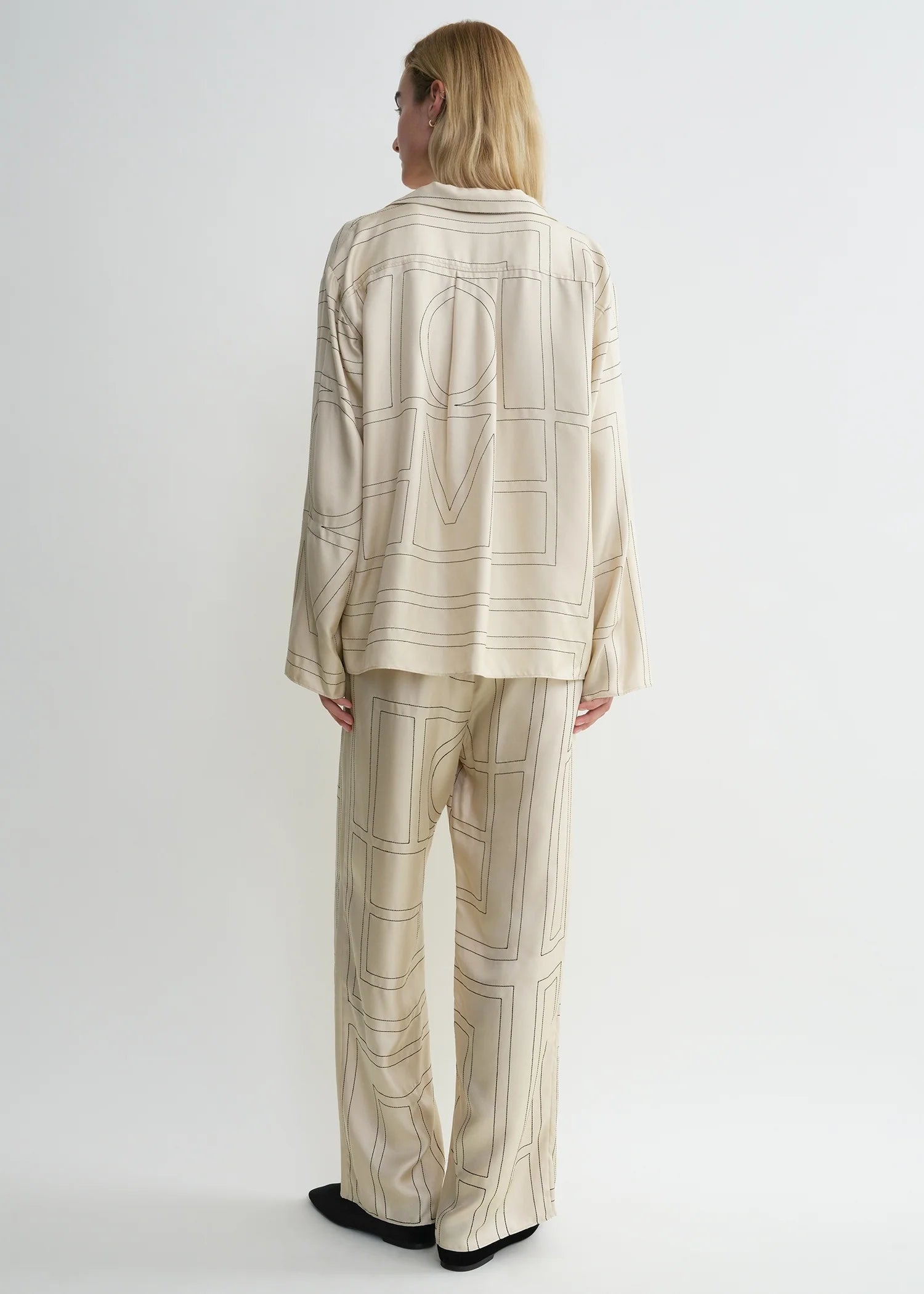 A person stands facing away, wearing the Toteme Monogram Silk PJ Top in a loose cream silhouette with a signature monogram pattern, set against a plain white background.