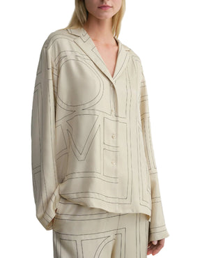 A woman wears the Toteme Monogram Silk PJ Top in beige with a bold monogram pattern. The image is cropped at her nose and above her knees.