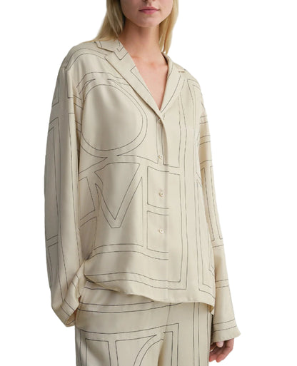 A woman wears the Toteme Monogram Silk PJ Top in beige with a bold monogram pattern. The image is cropped at her nose and above her knees.