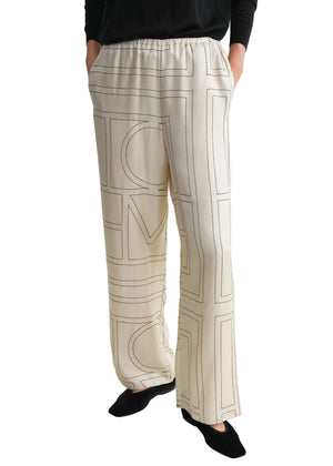 Someone wearing Toteme Monogram Silk PJ Bottoms—cream-colored trousers with black geometric line patterns—has hands in pockets and pairs them with a black top and black shoes.