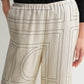 A person wears Toteme Monogram Silk PJ Bottoms featuring light tones and black geometric lines, paired with a black top and one hand in the pocket.