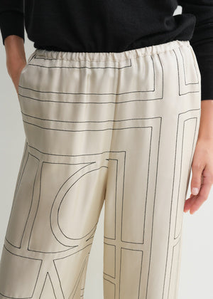 A person wears Toteme Monogram Silk PJ Bottoms featuring light tones and black geometric lines, paired with a black top and one hand in the pocket.