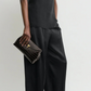 A person in a loose black sleeveless top and wide-leg trousers holds the Toteme T-Lock Embossed Croc Clutch against a plain background.