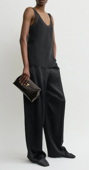 A person in a loose black sleeveless top and wide-leg trousers holds the Toteme T-Lock Embossed Croc Clutch against a plain background.