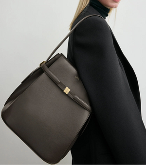 A person in a black blazer and dark turtleneck carries a Toteme Belted Bucket Bag made from brown Palmellato leather with gold accents over their shoulder.