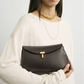 A woman holds a dark brown Toteme T-Lock Clutch, featuring Palmellato leather and a signature gold T-Lock clasp, while wearing a cream long-sleeve top and gold necklaces.