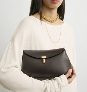 A woman holds a dark brown Toteme T-Lock Clutch, featuring Palmellato leather and a signature gold T-Lock clasp, while wearing a cream long-sleeve top and gold necklaces.