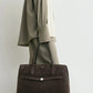 A person in loose taupe clothing with a cream sweater over their shoulders holds the Toteme Suede Lounge Tote, a large dark brown Italian suede bag with two handles.