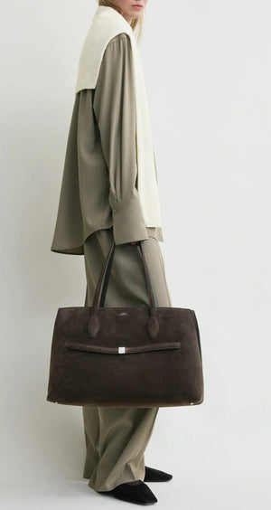 A person in loose taupe clothing with a cream sweater over their shoulders holds the Toteme Suede Lounge Tote, a large dark brown Italian suede bag with two handles.