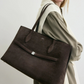 A woman in a taupe dress and cream sweater carries the Toteme Suede Lounge Tote—a spacious Italian suede bag perfect for effortless everyday style.