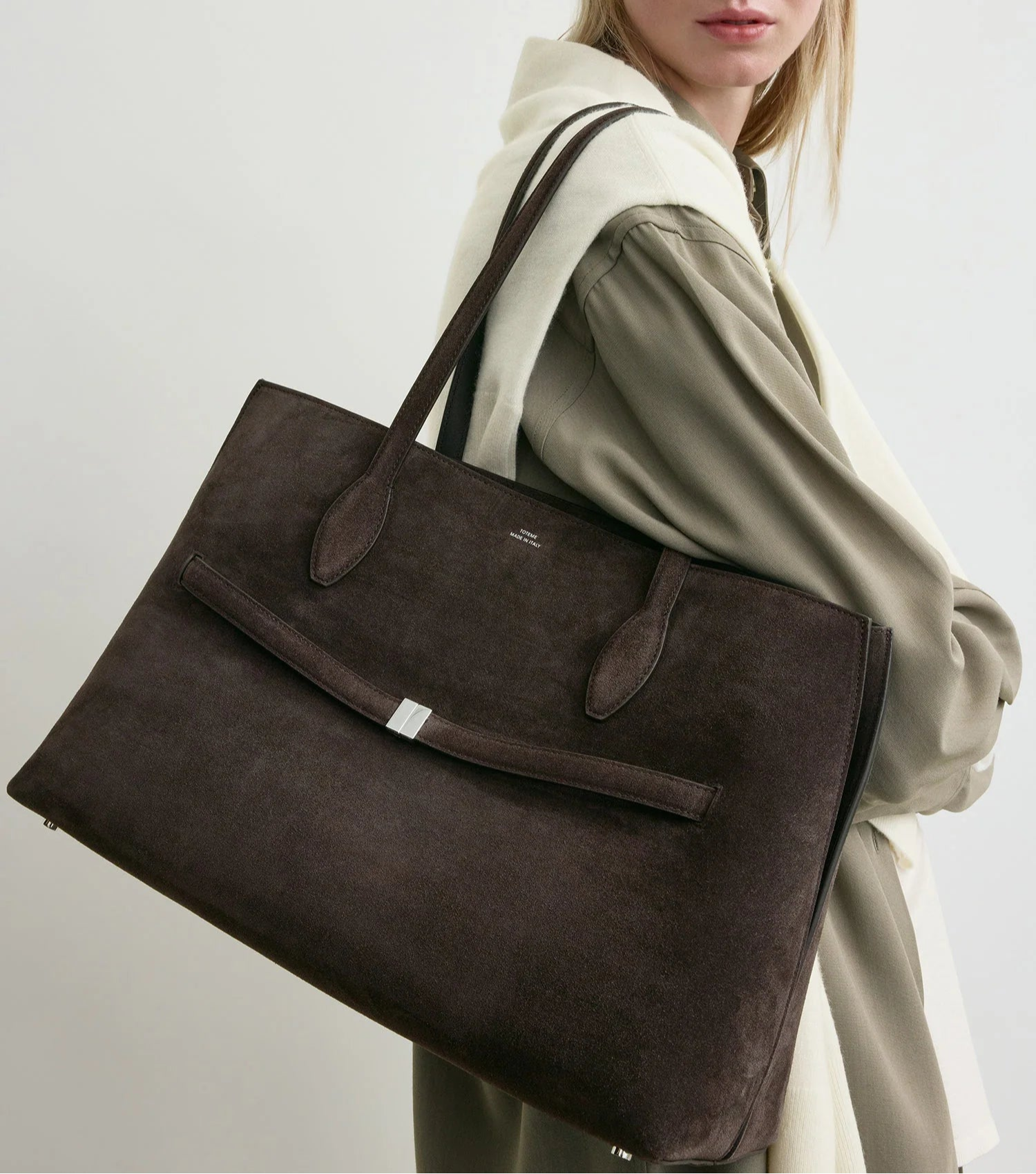 A woman in a taupe dress and cream sweater carries the Toteme Suede Lounge Tote—a spacious Italian suede bag perfect for effortless everyday style.