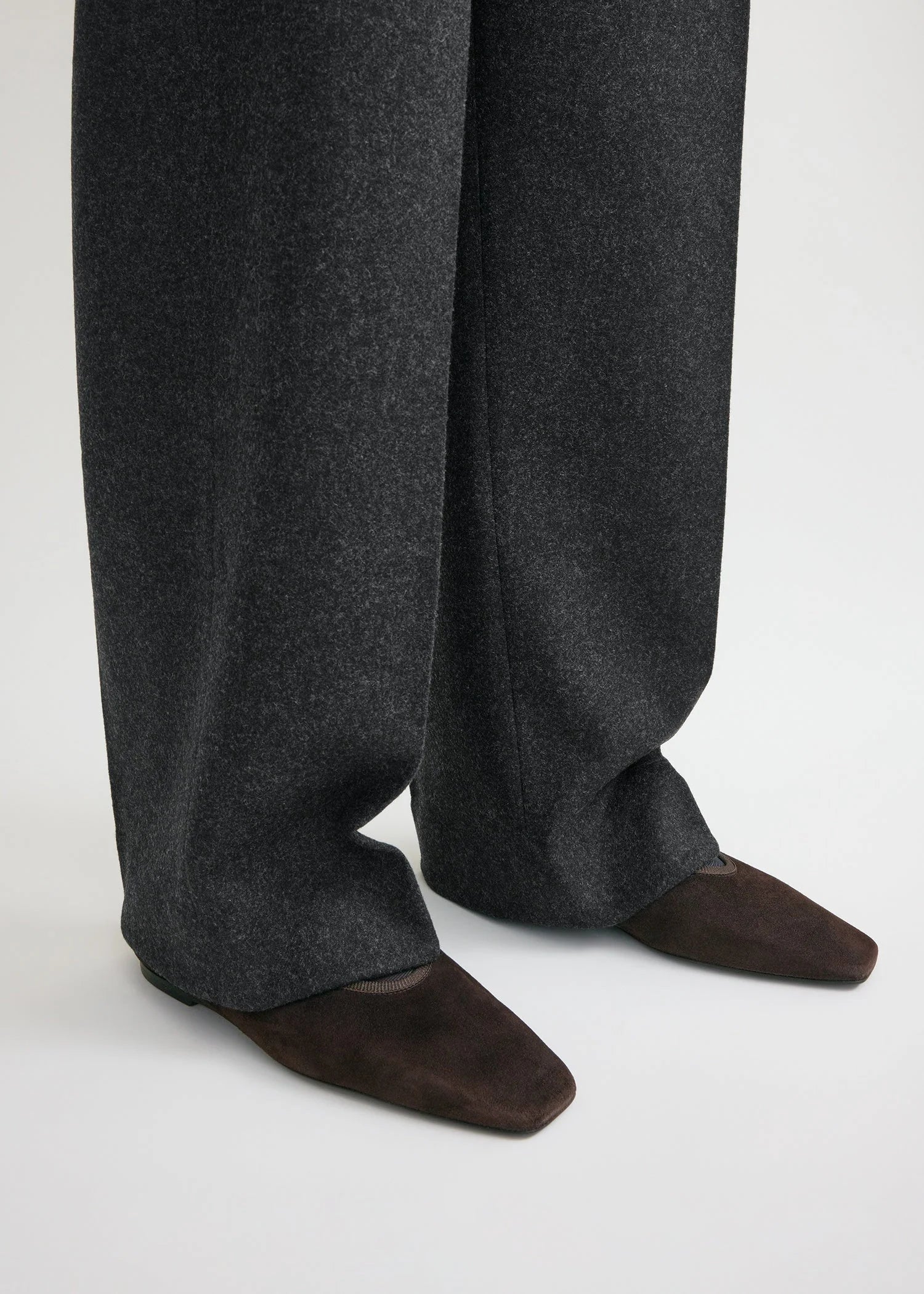 A person stands on a light-colored surface wearing dark gray wide-leg pants and Toteme’s Minimalist Suede Ballerinas, brown square-toe shoes made in Spain.