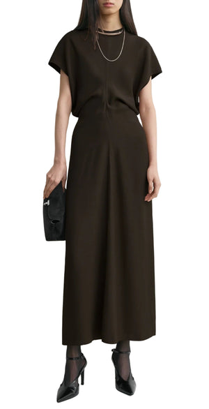 A woman wears Toteme's Slouch Waist Dress in black with short sleeves and a floor-length hem, styled with black heels, sheer socks, a necklace, and a black clutch.