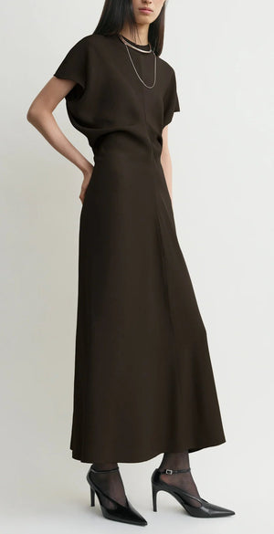 A woman stands against a plain light background wearing the Toteme Slouch Waist Dress—an ankle-length, short-sleeve black crepe dress—paired with black heels, sheer black tights, and a silver necklace.