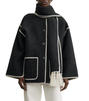 A person wearing Toteme's Embroidered Scarf Jacket—an oversized black wool-blend coat with white trim, large buttons, front pocket, and an attached matching fringed scarf—paired with white pants.