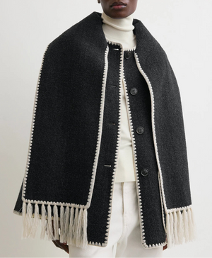A person wears the Toteme Embroidered Scarf Jacket—an oversized, wool-blend coat with white trim and fringed edges—layered over a white turtleneck and light-colored pants.