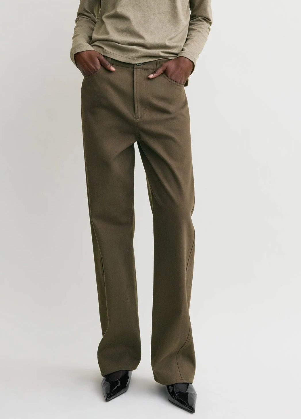 Wearing Toteme's Twisted Tailored Trousers—brown wide-leg style—paired with a beige long-sleeve top, this person stands hands in pockets, shown from shoulders down.