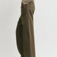 A person wearing Toteme's Twisted Tailored Trousers, styled with a beige long-sleeve top and black pointed shoes, stands against a plain light background.