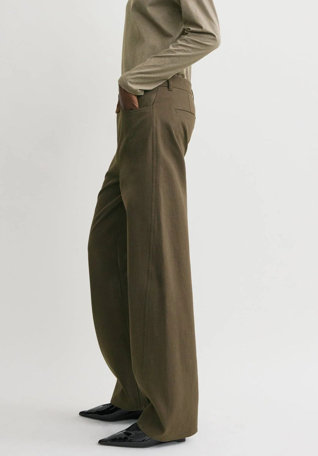 A person wearing Toteme's Twisted Tailored Trousers, styled with a beige long-sleeve top and black pointed shoes, stands against a plain light background.